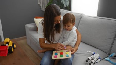 Genç oğluyla kanepede oynayan bir kadın, oyuncaklarla çevrili, sıcak bir oturma odasında aile sevgisini ve yakınlaşmayı vurguluyor..