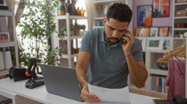 Delikanlı, dekoratif unsurlarla çevrili bir mağazada telefonla konuşurken kağıtları gözden geçiriyor ve bir dizüstü bilgisayarla çalışıyor.