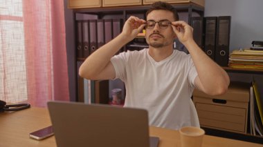 Yakışıklı, sakallı bir İspanyol, arka planda dosyalar, dizüstü bilgisayarlar, kahve bardakları ve pembe perdelerle bir ofiste otururken gözlüklerini ayarlıyor..