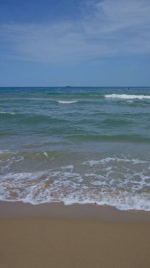 Sade bir deniz manzarası, yumuşak dalgalar sergiliyor kumlu bir kumsalda, açık mavi gökyüzü ile..