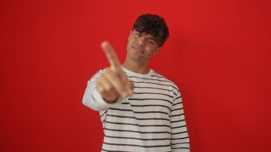 Yakışıklı bir İspanyol genç, izole edilmiş kırmızı bir arkaplanda kamerayı işaret ederek güven ve bağlantıyı ifade ediyor..