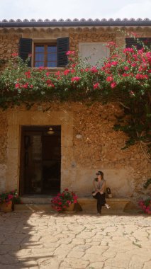 İspanya, Mallorca 'da, renkli çiçeklerle dolu taş bir evin önünde, huzurlu ve güneşli bir yaz ortamı sergileyen genç bir kadın..