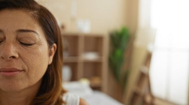 Yumuşak ışık ve sakinleştirici dekorla dolu sakin bir spa odasında dinlenen, gözleri kapalı, olgun İspanyol bir kadın.