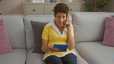 Kısa saçlı, yaşlı İspanyol bir kadın bir kanepede oturuyor, telefonda konuşurken tablet kullanıyor, rahat bir ev ortamında..