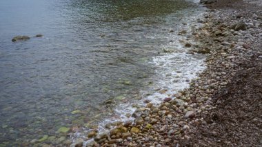 Sakin su dalgaları yumuşak renkli çakıl taşları ve taşların üzerinde yavaşça yuvarlanan kayalık kıyısı açık havada huzurlu bir sahneyi gözler önüne seriyor..
