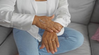 Orta yaşlı bir kadın evdeki rahat bir oturma odasında otururken ellerine krem sürüyor. Kendi kendine bakım ve ev içi rahatlığı vurguluyor..