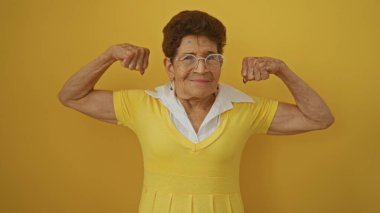 Kısa saçlı yaşlı İspanyol kadın izole edilmiş sarı bir arka planda kaslarını esnetiyor. Güç ve kendine güven gösteriyor..