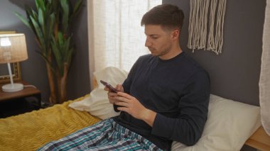 Genç bir İspanyol adam, rahat bir yatak odasında, rahat bir ortamda yastığa yaslanırken telefonunu kullanarak yatağında dinleniyor..