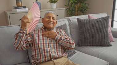 Hispanic elderly man fanning himself and smiling in a cozy living room setting, embodying warmth and comfort. clipart