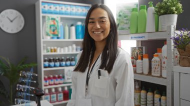 Eczanede beyaz önlüklü ve isim etiketli çeşitli ürünlerin raflarının önünde gülümseyen genç bir kadın.