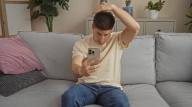 Yakışıklı İspanyol bir adam oturma odasında bir kanepede oturuyor, telefonuna bakıyor ve saçına dokunuyor..