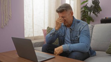 Orta yaşlı bir adam oturma odasında stresli ya da hayal kırıklığına uğramış hissediyor, masadaki bir dizüstü bilgisayarla evdeki koltukta oturuyor..