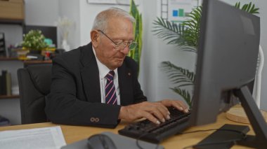 Yaşlı, gri saçlı, beyaz bir adam modern bir ofis ortamında masaüstü bilgisayarında kitap rafları ve arka planda bitkilerle yazı yazıyor..