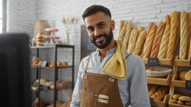 Yakışıklı, sakallı, önlüklü, fırında dikilip arka planda ekmek raflarıyla kameraya bakan bir adam.