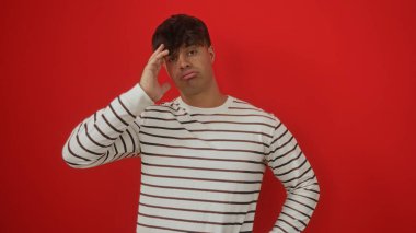 Handsome young hispanic man in a striped shirt posing against a vibrant red wall, conveying a thoughtful expression. clipart