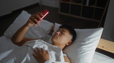 Genç adam yatakta uzanıyor, akıllı telefon kullanıyor, evdeki bir yatak odasında, modern yaşam tarzı ve teknolojisine odaklanmış, Çin 'de rahat ve rahat bir ev dekorunu sergiliyor.