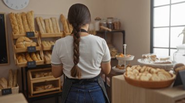 Esmer saçlı genç bir kadın pastanede çalışıyor ve çeşitli ekmek ve hamur işleri sergileniyor.