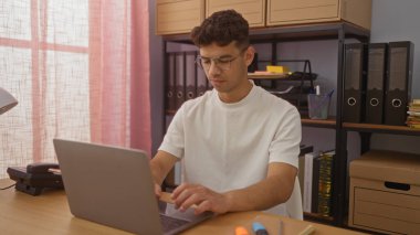 Düzenli bir ofis ortamında dizüstü bilgisayarda çalışan yakışıklı İspanyol bir adam. Arkasında dosya ve kutularla dolu raflar var..