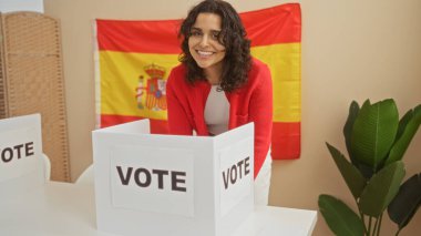 A young, attractive hispanic woman stands smiling in front of a voting booth with a spanish flag in the background, indoors. clipart