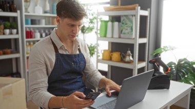 Ev dekorasyon mağazasında akıllı telefon ve dizüstü bilgisayar kullanan önlüklü genç adam, çeşitli dekoratif eşyalar ve bitkilerle çevrili..
