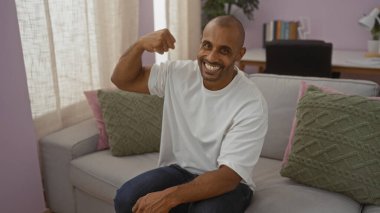 Yakışıklı genç adam kapalı bir salonda kanepede otururken kolunu bükerek gülümsüyor.