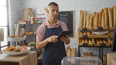 Fırında elinde tablet olan, önlük takan, rafları ekmek ve hamur işleriyle dolu genç bir adam..