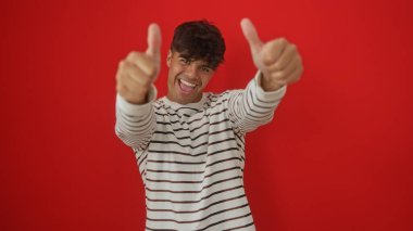 Çizgili tişörtlü genç İspanyol adam, neşeli kırmızı bir arka planda başparmağını kaldırıyor ve gülümsüyor..