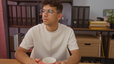 Ofiste oturan gözlüklü genç İspanyol bir adam. Elinde bir bardak var. Etrafı kitap ve kitaplarla çevrili..