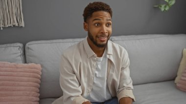 Genç adam sıcak bir oturma odasında oturuyor, sıcak, dostane bir ifadeyle, modern dekorla çevrili.