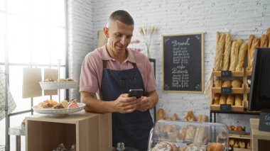 Pastanedeki genç adam tezgahın arkasında taze ekmek ve hamur işleriyle dikilirken telefonunu kontrol ediyor.