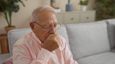 Oturma odasında oturan gri saçlı, gözlüklü ve pembe gömlekli yaşlı bir adam, rahat bir evde düşünceli bir yüz ifadesi sergiliyor..