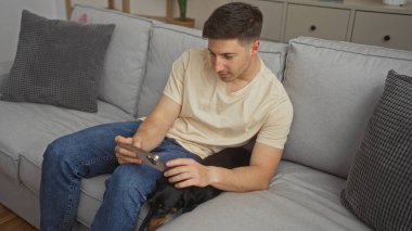 Genç bir İspanyol adam oturma odasında kanepede oturuyor. Tekel köpeği bacağında dinlenirken akıllı telefonunu kullanıyor..