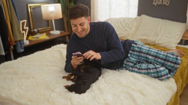 Genç bir İspanyol adam yatak odasında bir yatakta uzanıp telefonuna bakarken daksund köpeğiyle oynuyor..