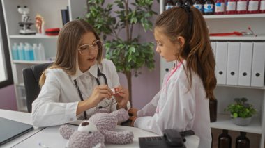 Klinikteki kadın doktor, çocukluk hastası olarak bir kızın elini kontrol ediyor. Hastane odasında tıbbi aletlerle ve oyuncak ayıyla dolu bir masada oturuyor..