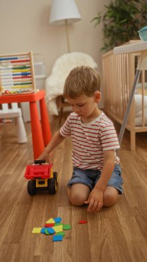 Sevimli sarışın çocuk, oyuncak bir traktörle yerde oynuyor. Kapalı bir anaokulu ortamında. Renkli bloklar etrafa saçılmış..