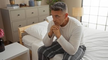 Yatak odasında yakışıklı, orta yaşlı bir adam. Düşünceli bir şekilde yatakta oturuyor.
