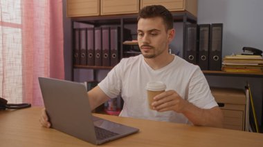 Ofiste elinde kahve fincanı tutarken dizüstü bilgisayarla çalışan yakışıklı bir İspanyol..