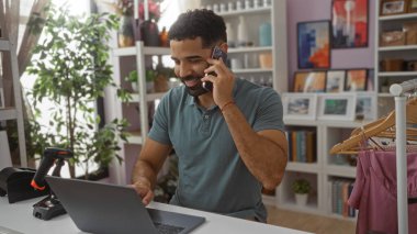 Dizüstü bilgisayarda çalışan ve dekore edilmiş bir ev dekoratöründe telefonla konuşan genç bir adam. Arka planda kitap rafları, giyim rafları ve bitkiler var..