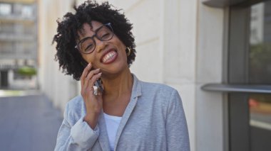 Kıvırcık saçlı güzel bir Afro-Amerikalı kadın şehir caddesinde telefonda gülümsüyor ve konuşuyor..