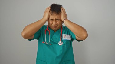 Tıbbi önlüklü genç bir Çinli steteskoplu bir adam stresli görünüyor, izole edilmiş beyaz bir arka plan yüzünden kafasını tutamıyor..