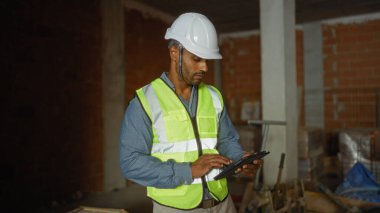 İnşaat alanında güvenlik kaskı ve yüksek görüş mesafesi olan yelek giyen genç adam tablet kullanıyor.