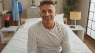 Yakışıklı orta yaşlı bir adam evdeki rahat bir yatak odasında bir yatakta oturuyor, gülümsüyor ve beyaz bir gömlek giyiyor, arka planda bitkiler ve mobilyalar var.