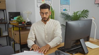 Modern ofis ortamında bilgisayar başında çalışan İspanyol bir adam. Etrafta kitaplar ve bitkiler var..