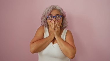 Gri saçlı kadın, pembe bir arka plan ve beyaz kolsuz bir bluz giyerek ağzını sürpriz bir şekilde kapatıyor.