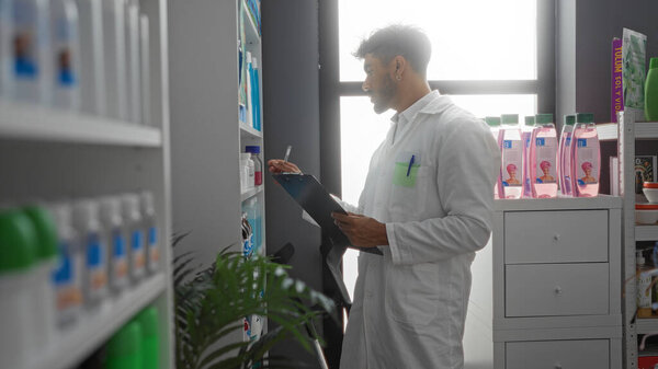 Young hispanic man working in a pharmacy, wearing a white coat and holding a clipboard, checking stock of various health products on shelves.