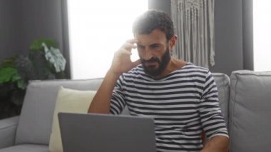 Çizgili tişörtlü, sakallı genç adam oturma odasında oturmuş endişeli bir ifadeyle dizüstü bilgisayar kullanıyor.