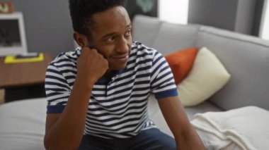 Evdeki kanepede oturan genç adam, rahat bir oturma odasında yumuşak yastıklar ve modern dekorlarla çevrili, sıradan çizgili gömleğiyle düşünceli bir şekilde gülümsüyor..