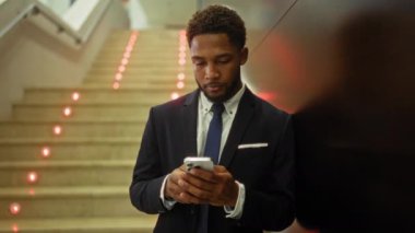 Takım elbiseli Afro-Amerikan genç adam kapalı bir iş yerinde akıllı telefon kullanıyor ve arka planda ışıklı merdivenler var.
