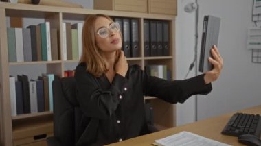 Ofisinde tabletli selfie çeken bir kadın. Etrafı kitaplık ve belgelerle çevrili. Gözlüklü ve siyah gömlekli.