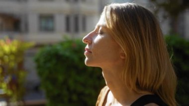 Şehir parkında gün ışığından hoşlanan kadın yeşil ve şehir manzarasıyla çevrili huzurlu ifadesini vurguluyor..